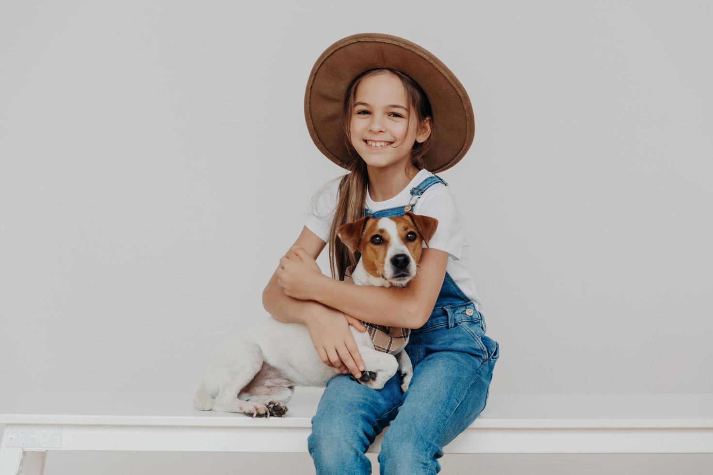 Indoor shot of adorable small preschool girl dressed in fashionable outfit, huggs jack russel terrier dog, plays with best animal friend, love each other. Children, pets and friendship concept photo