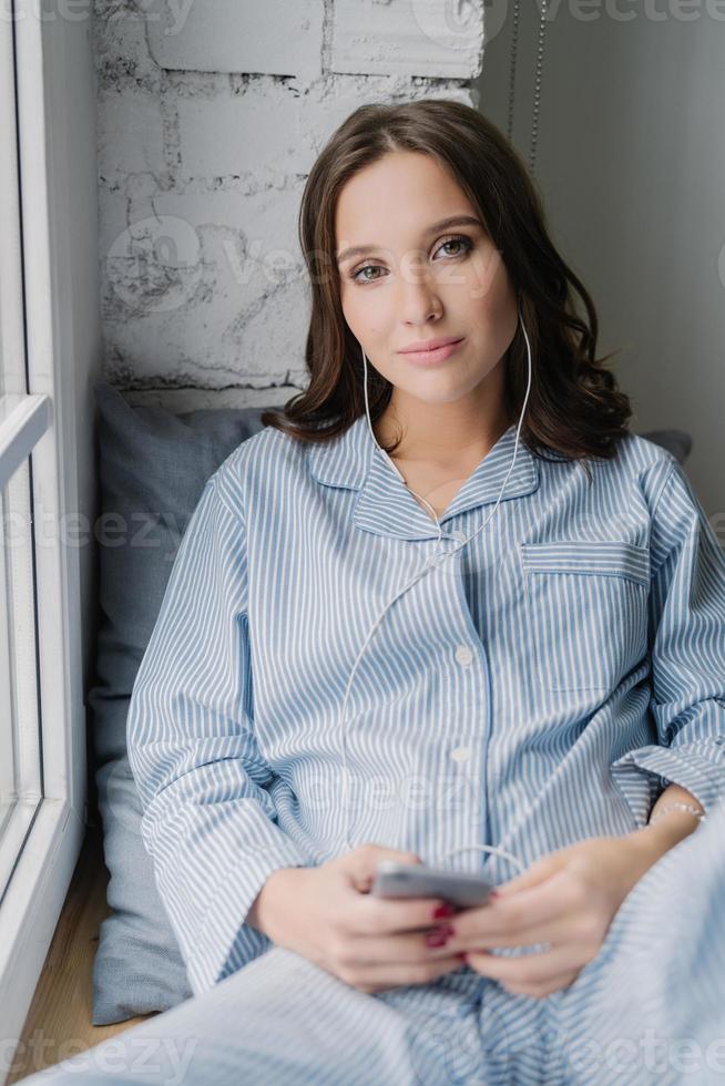 Female blogger in domestic clothes listens music in earphones and chats via cellular while sits on window sill, has calm look expression, looks seriously directly at camera, poses at home or apartment photo