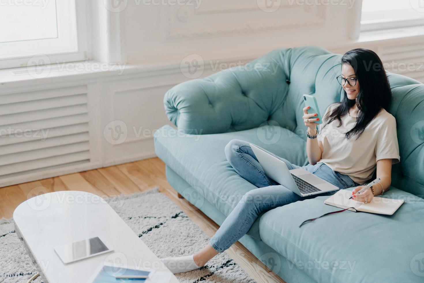 Above shot of pretty brunette woman focused in samrtphone display, writes down necessary information in notebook, watches training webinar, poses in cozy room on comfortable couch, works from home photo