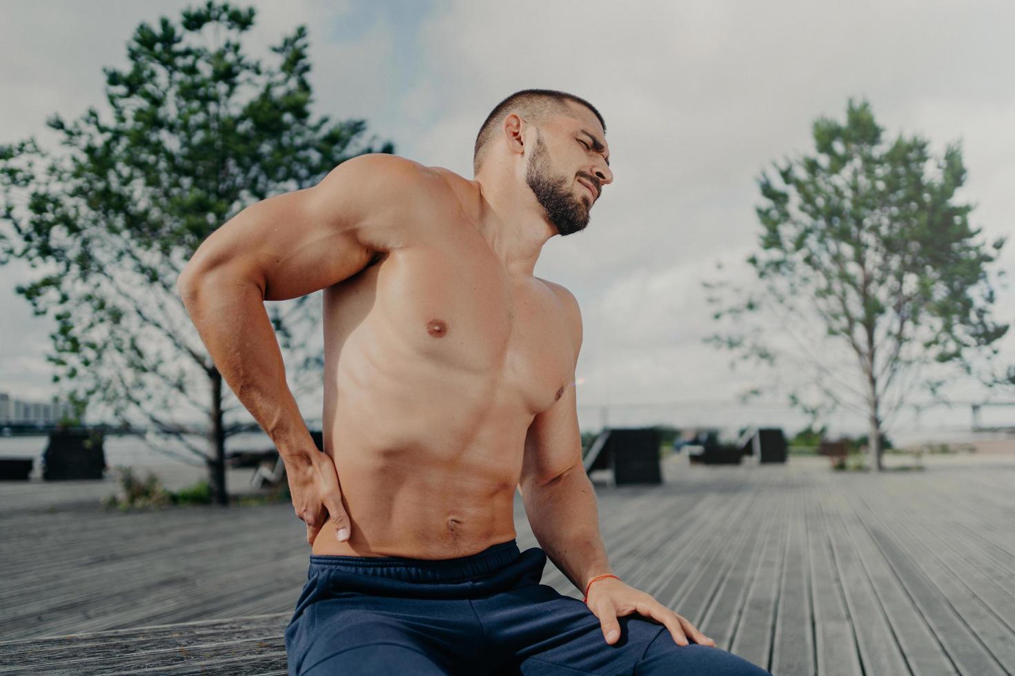 Displeased unshaven European sportsman touches lower back and feels backache after physical training exercises, poses outdoor. Sport exercising injury. Athletic guy suffers pain, needs massage photo