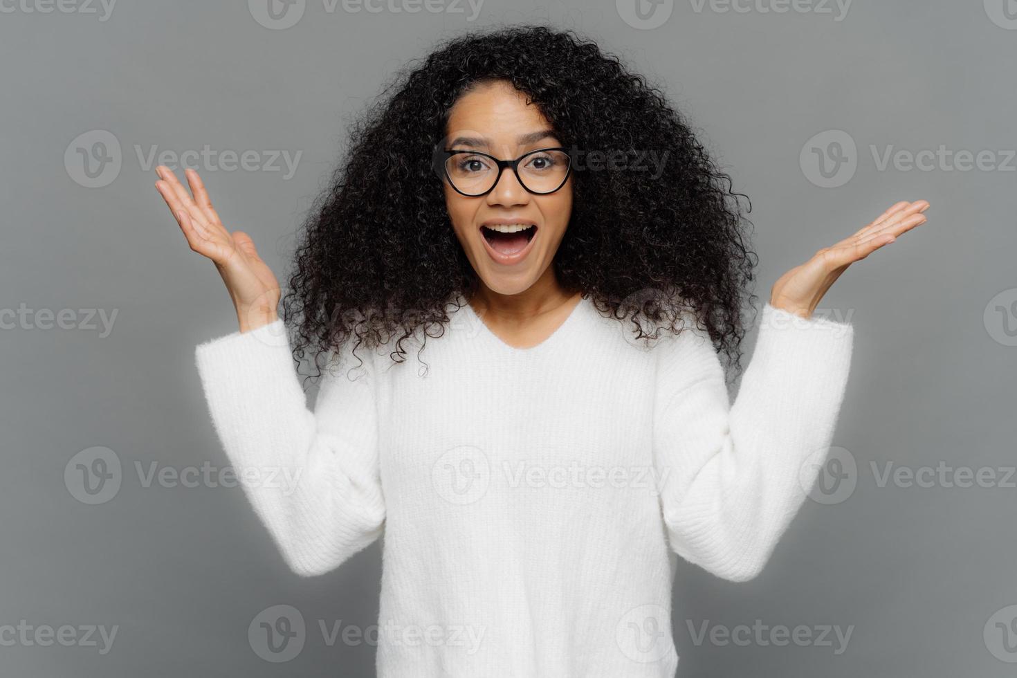 Satisfied African American woman raises hands in eureka gesture, keeps palms raised, wide opened mouth, wears optical eyewear and white jumper, isolated over grey background. Wow, its awesome. photo