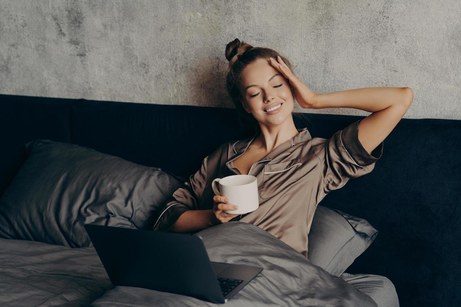 Charming relaxed young woman lying in bed with laptop at home and drinking coffee photo