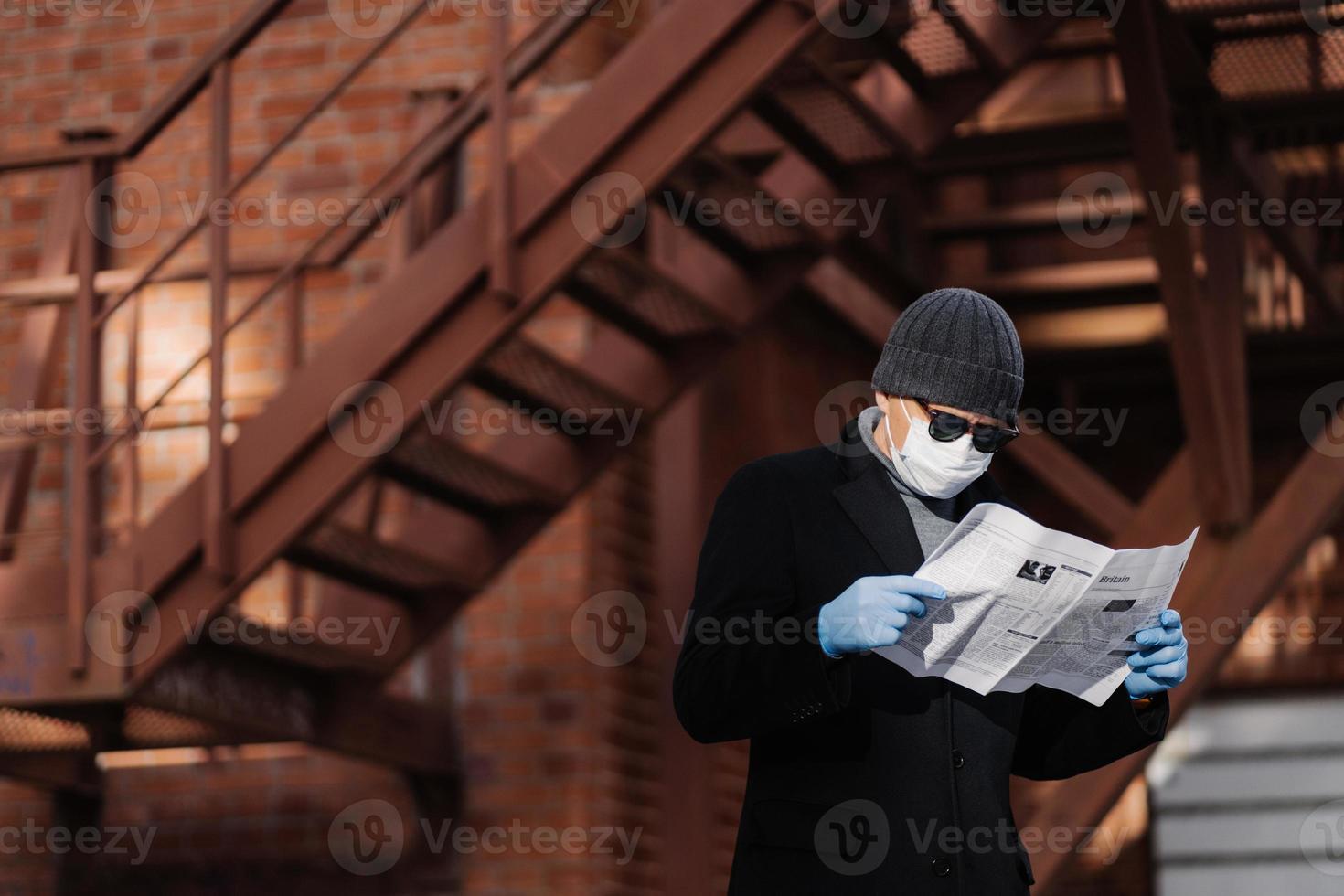 Coronavirus precautions. Male model avoids crowded places, fights against new virus, reads attentively article about covid-19 symptoms, holds newspaper, wears protective face mask and medical gloves photo