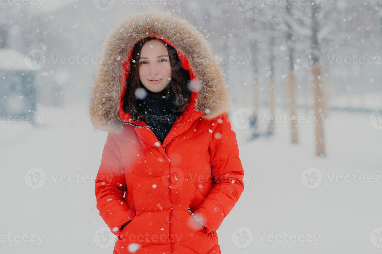 Lovely female model in red jacket, stads outdoor during snowfall, looks with dark eyes at camera, going to have stroll with boyfriend, breathes fresh air. People, winter time and free time concept photo