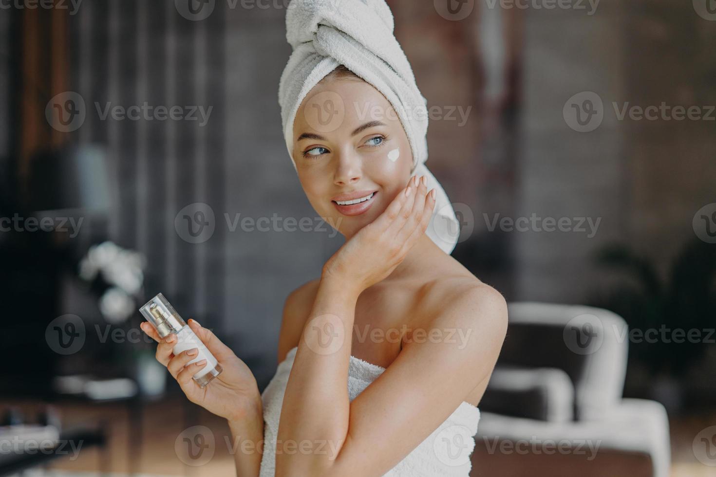 Indoor shot of young smiling woman applies moisturizer cream on face, takes care of her skin and complexion, puts lotions, has minimal makeup, wrapped in bath towel. Beauty, cosmetology concept photo