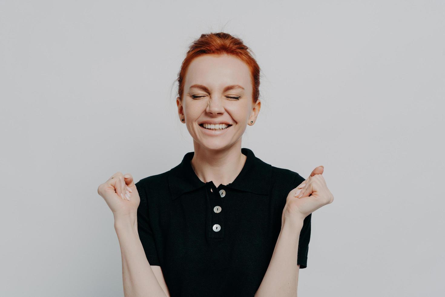 Happy euphoric red haired female with closed eyes holding clenched fists up and celebrating win photo