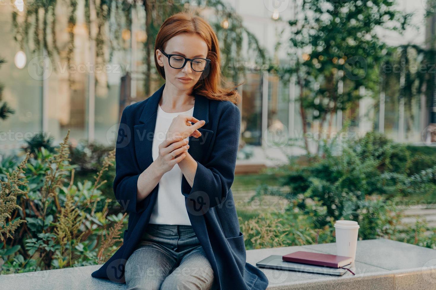 Young woman suffers from pain in wrist sits outside on bench focused down photo