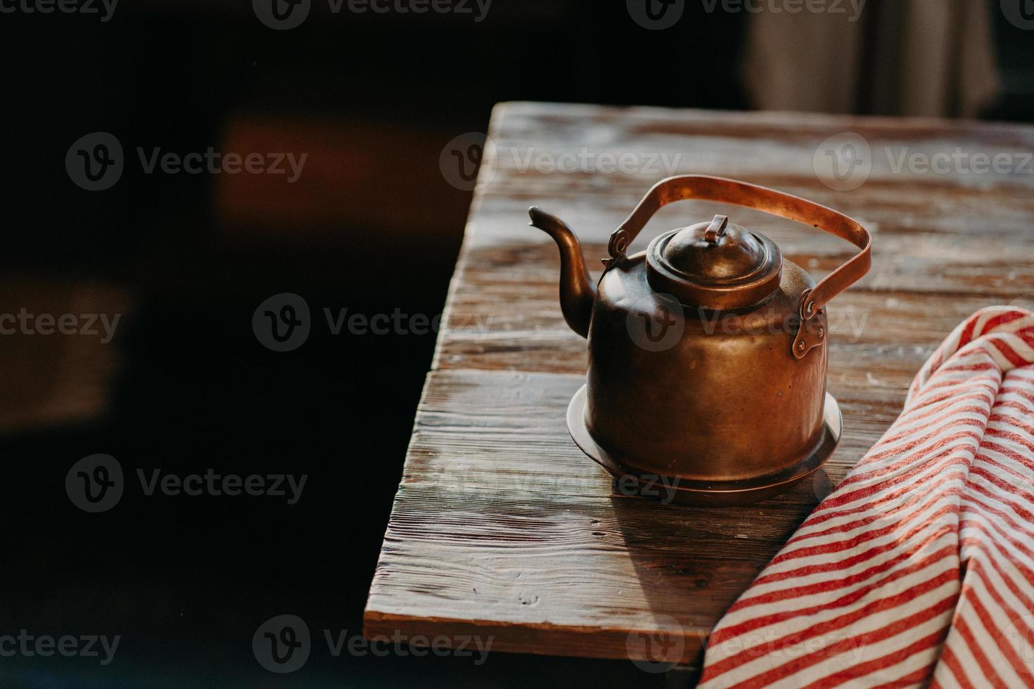 Old copper metal teapot on wooden table in dark room. Red striped towel nearby. Antique kettle for making tea or coffee. Cooking equipment photo