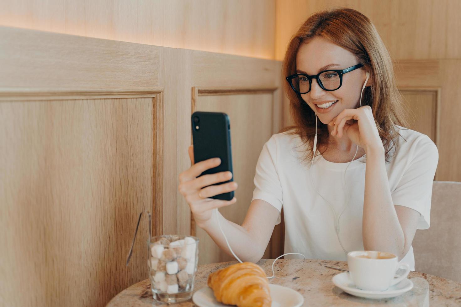 toma interior de una joven pelirroja alegre que usa teléfonos móviles y auriculares para conversar en línea foto