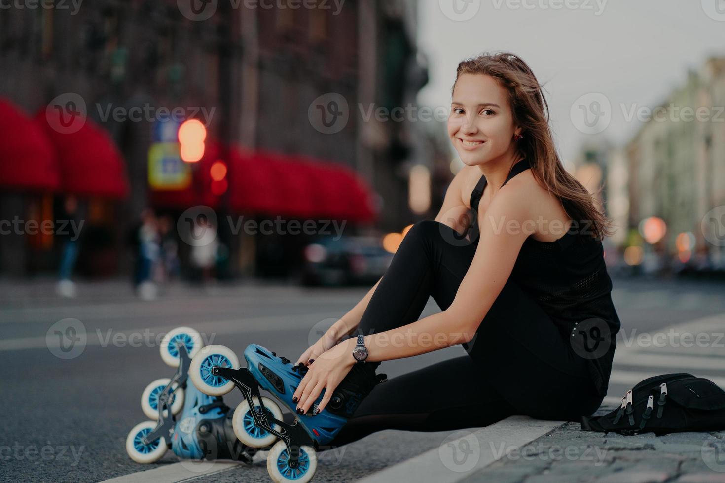 Fit positive woman puts on rollerblades sits on asphalt against blurred street background has positive expression goes in for sport has outdoor fitness activities during summer time. Hobby concept photo