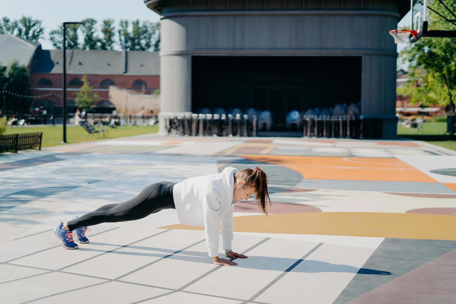 Full length side view shot of young active woman stands in plank position dressed in sweatshirt leggings sneakers has training outdoors on stadium goes in for sport. Morning workout concept. photo