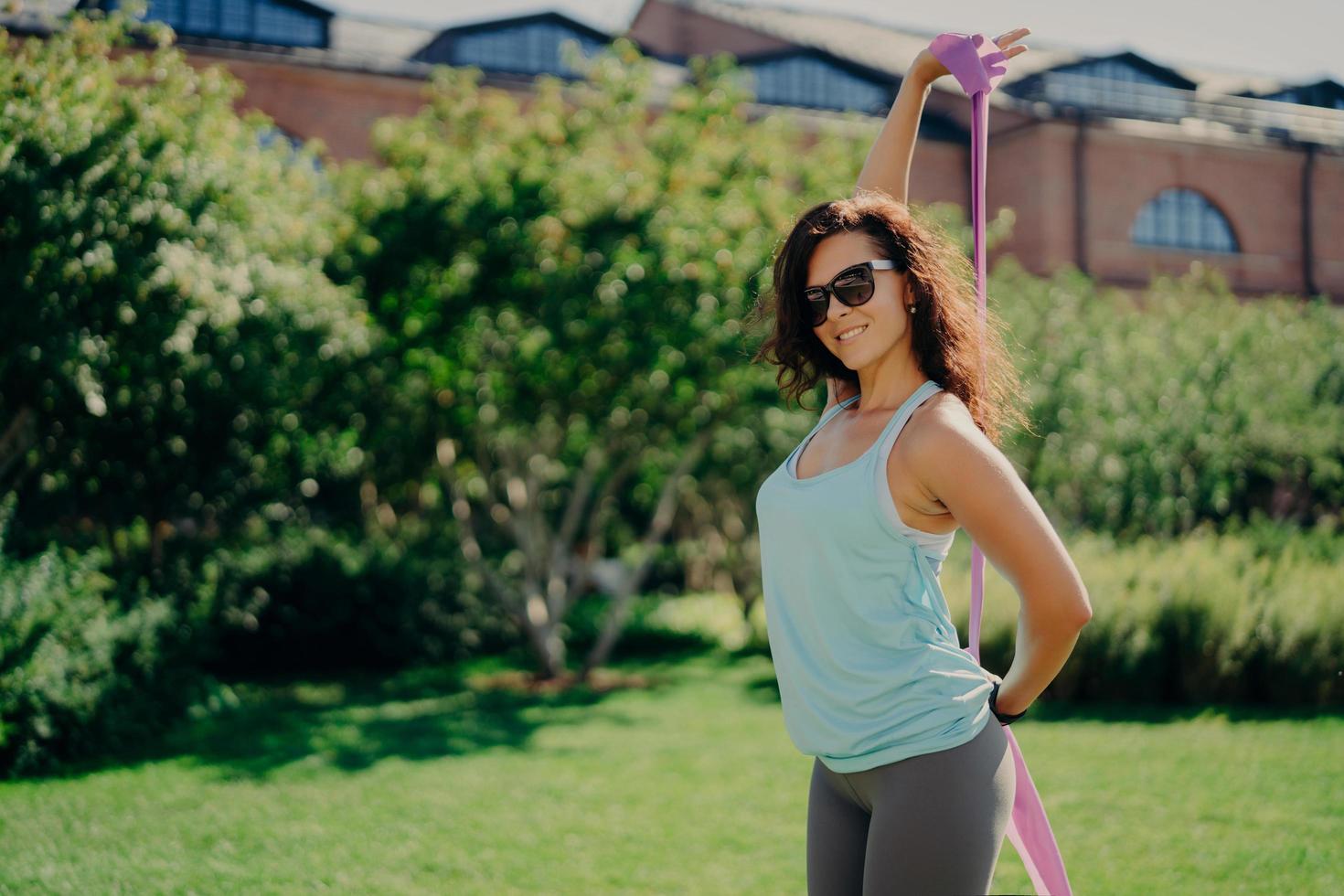 Fitness athletic woman does workout with rubber band dressed in sportsclothes trendy sunglasses poses outside happy against green nature background. Gymnastics coach female shows exercises outdoor photo