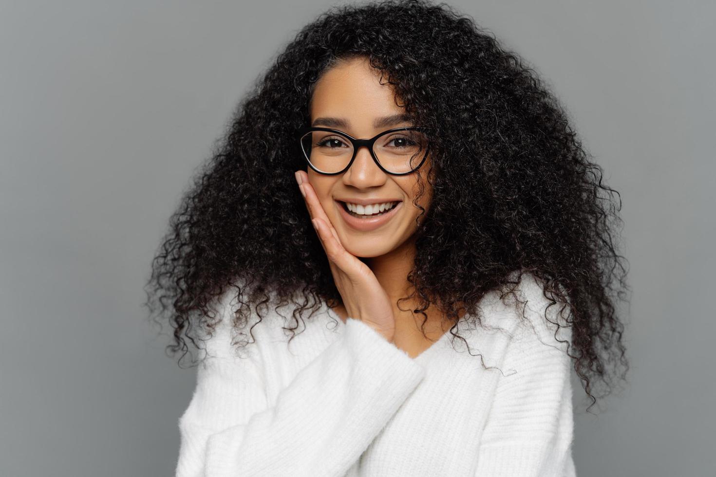 Portrait of good looking Afro woman touches cheek, smiles happily, enjoys receiving compliment, feels shy, dressed in white casual sweater, poses indoor. People, positive emotions and feelings concept photo