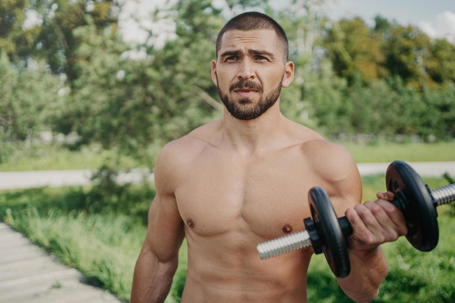 Outdoor shot of muscular bodybuilder with naked torso raises barbell and has strong biceps workout in open air looks thoughtfully into distance uses sport equipment. Handsome athlete with dumbbell photo