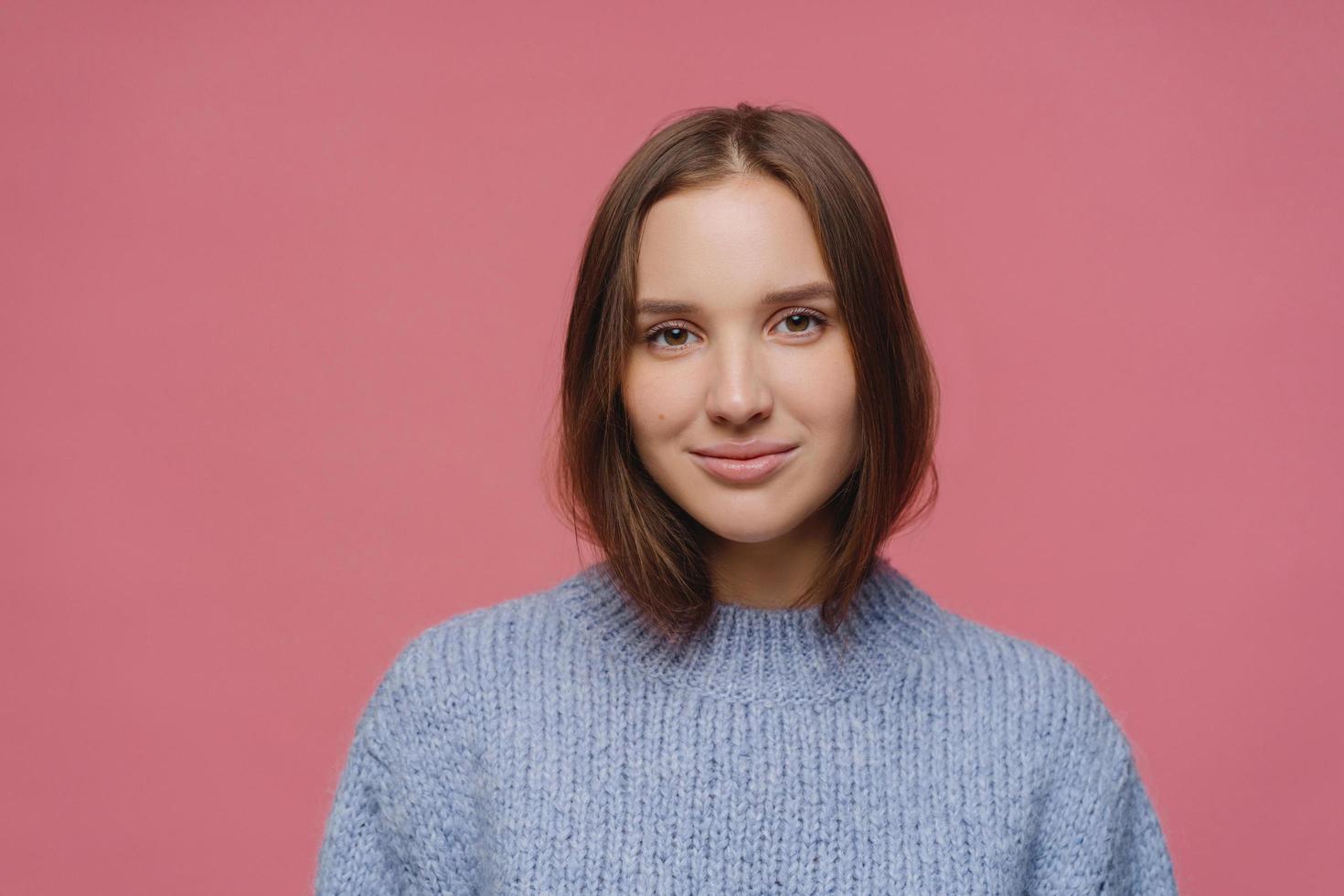Beautiful adult woman with dark hair, has calm face expression, wears knitted sweater, poses at studio against pink background, thinks about something pleasant during winter time, has tender look photo