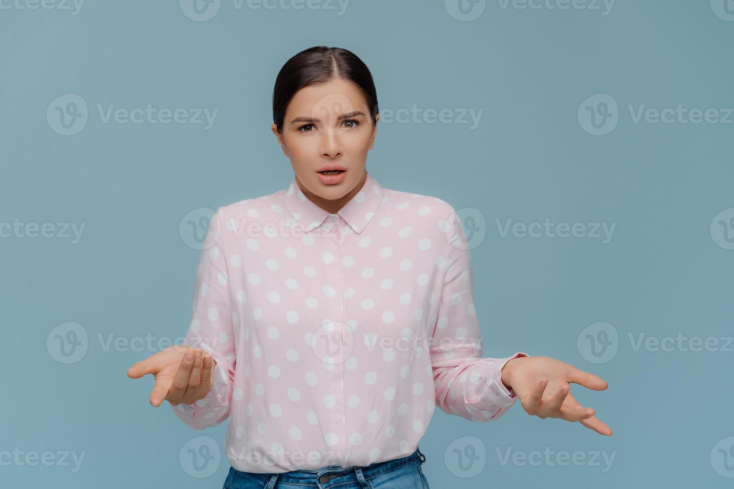 Pretty dark haired woman with makeup, spreads hands and looks doubtfully, feels misunderstanding, wears polka dot shirt and jeans, cannot make decision, poses indoor over blue studio background photo