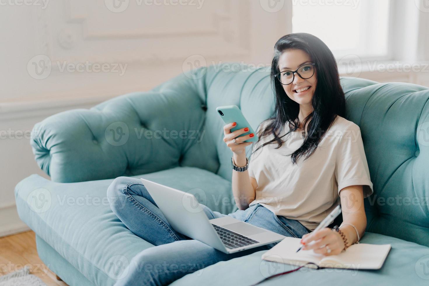 Glad female student works on course paper, surrounded with modern gadget, makes notes in notepad, feels relaxed at comfortable sofa, works online, wears casual clothes, watches video online. photo