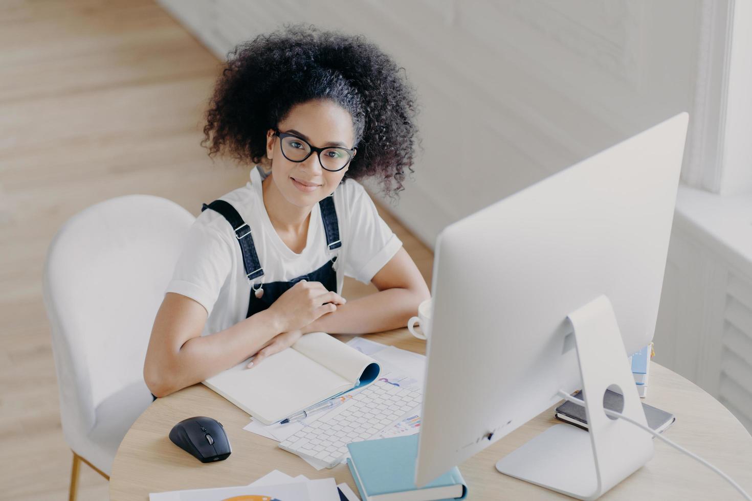 Top view of delighted curly female worker poses at desktop, dressed in casual wear, works on computer, makes financial report. Afro American student prepares for final exam in coworking space photo