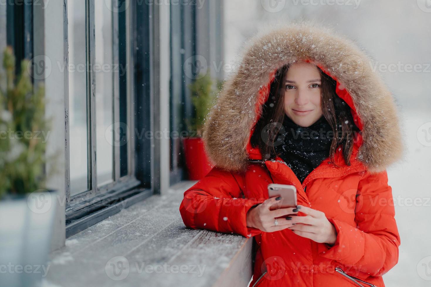 Smiling young European female in red coat, has black manicure, uses modern electronic gadget for communication in internet with friends, likes winter weather, sends photo in social networks.