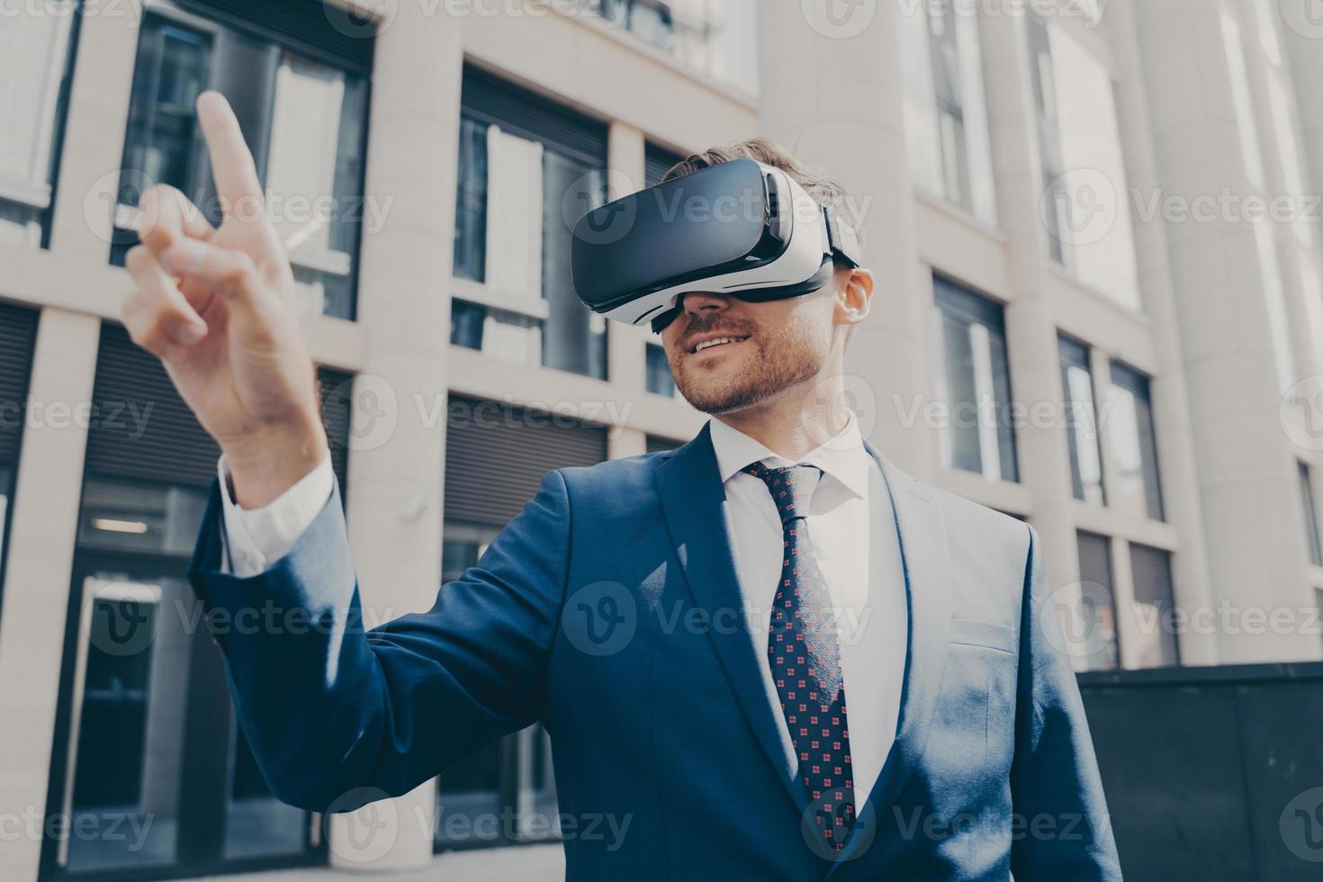 Cheerful businessman trying to touch something in virtual reality, standing near office building photo