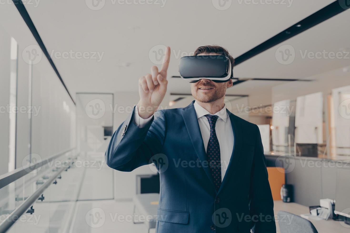 Smiling male entrepreneur using vr goggles for 3d visualization, pointing with finger photo