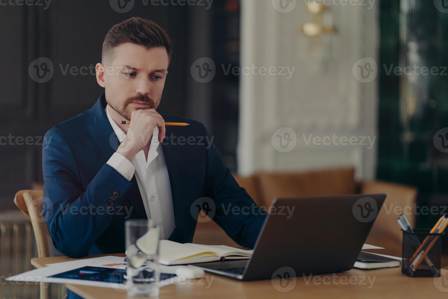 Serious male entrepreneur holds chin makes notes in diary works on laptop computer makes money online dressed formally writes information. Confident managing director involved in working process photo