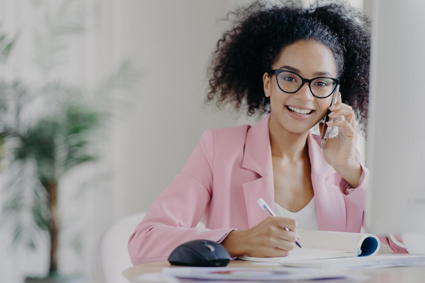 Happy African American woman with curly hairstyle, writes some information, speaks via cell phone, sits at desk against blurred office background. People, technology, communication, business concept photo