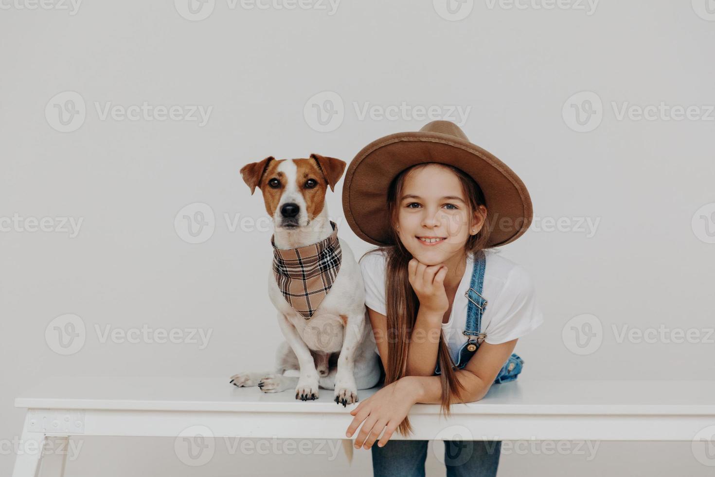 Content pretty girl wears hat, leans at white table, poses near pedigree dog, enjoy spending free time together, going to have walk, rest at home. Children, happiness, animals, lifestyle concept photo