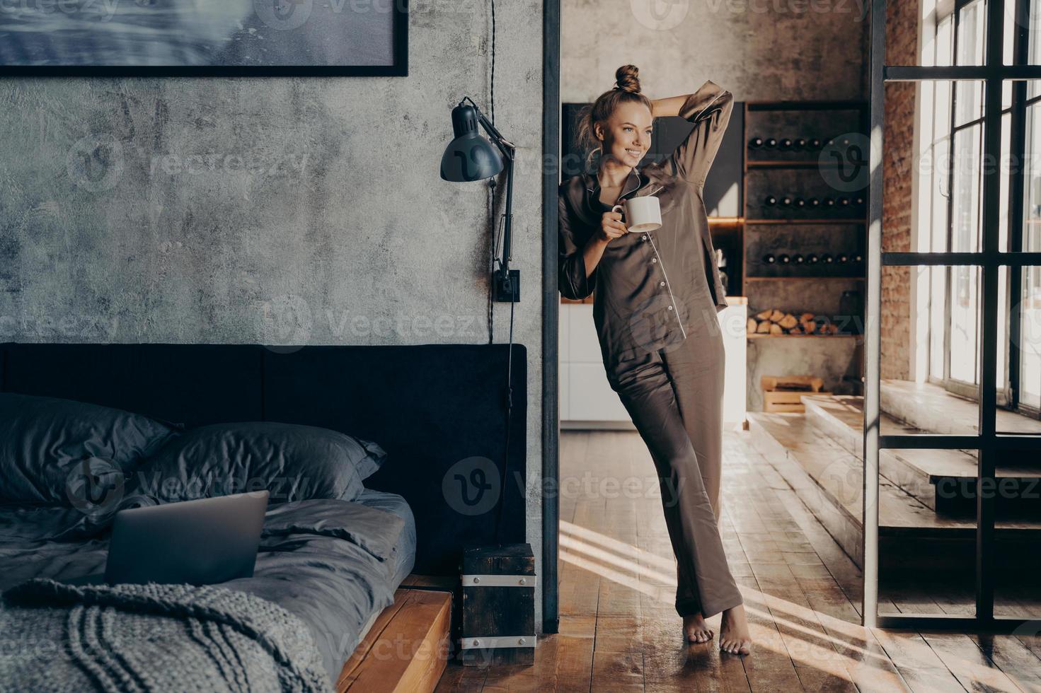 joven mujer bastante delgada con pijama de satén marrón sosteniendo una taza de café mientras está de pie en el dormitorio foto