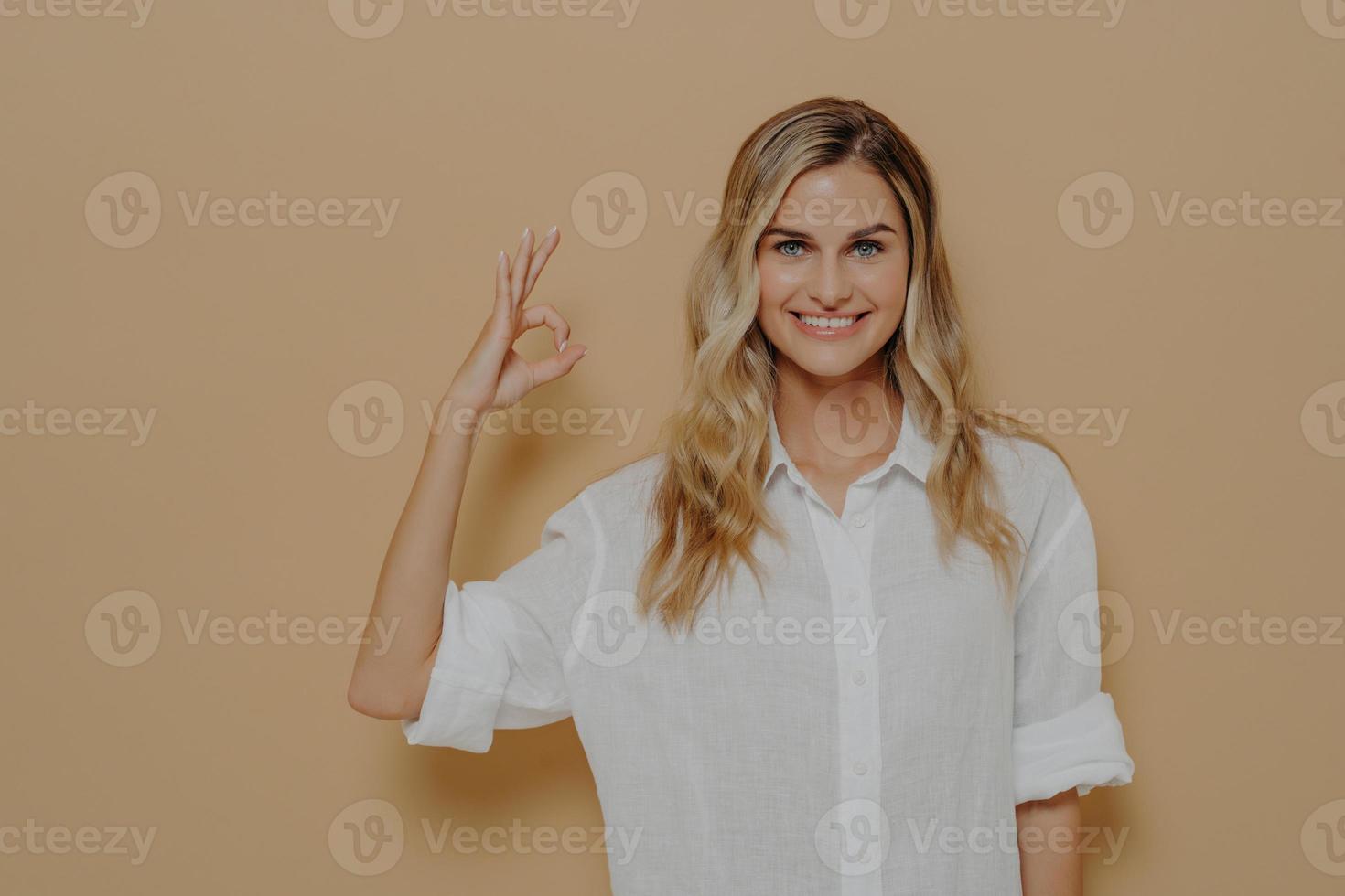 hermosa joven rubia con camisa blanca sonriendo positivamente haciendo un buen gesto con la mano y los dedos foto