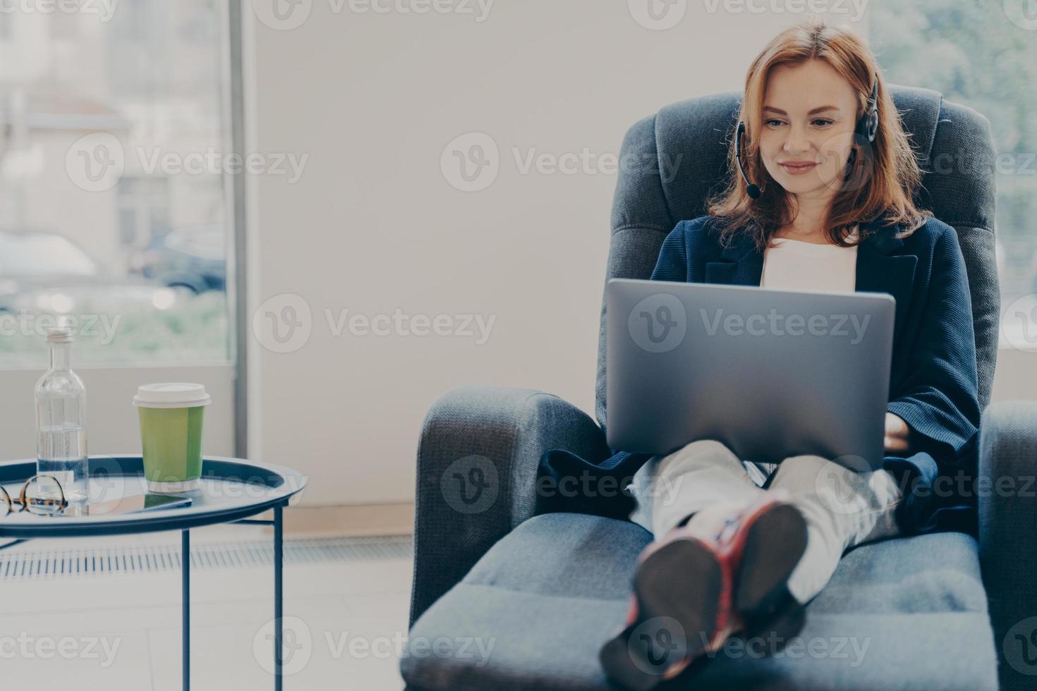 Smiling red-haired woman working in headphones on laptop computer in modern open plan office photo