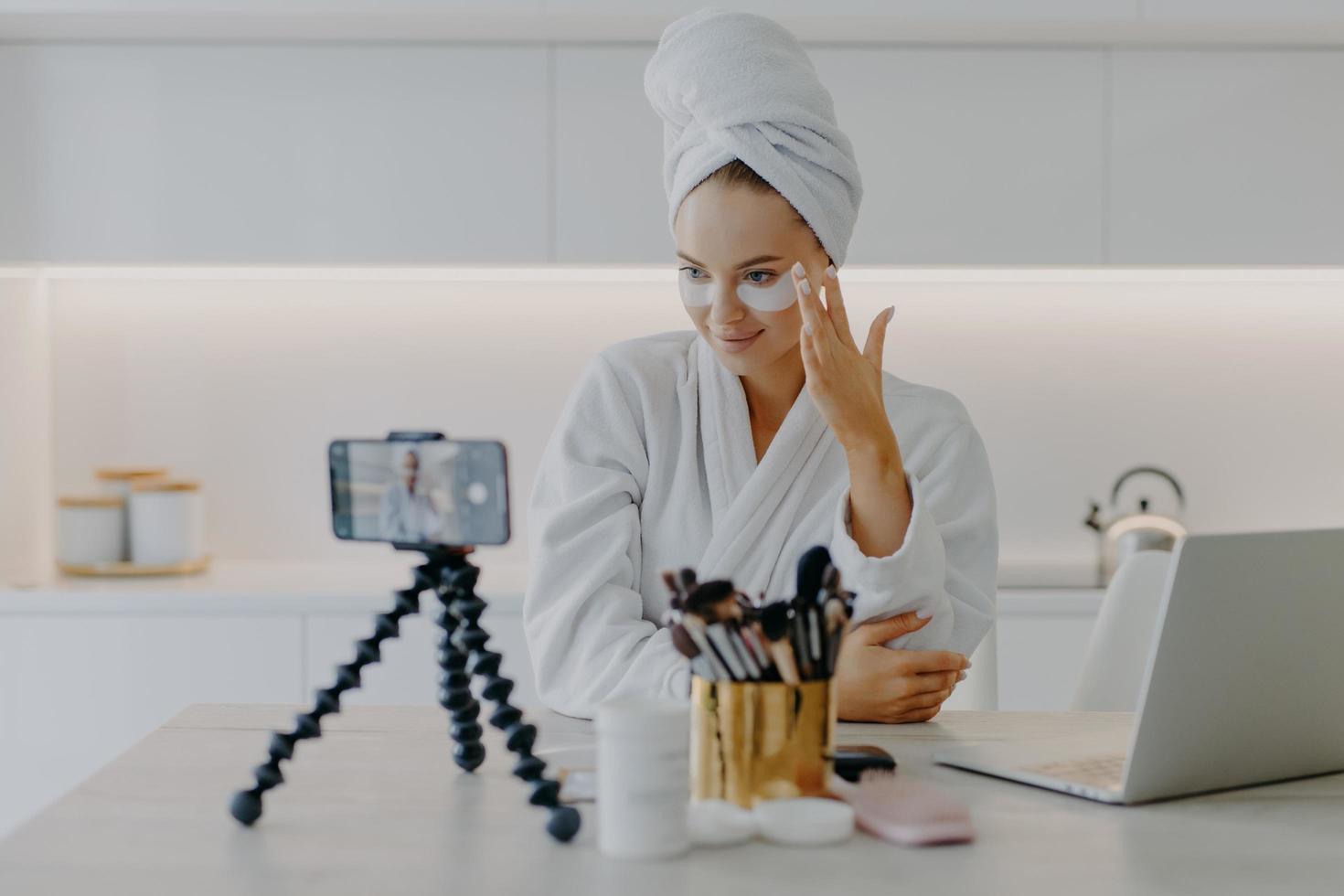 Lovely young female model advertises for internet views cosmetic product cares about skin in under eye area applies hydrogel patches records video on smartphone device for her blog. Vlogging concept photo