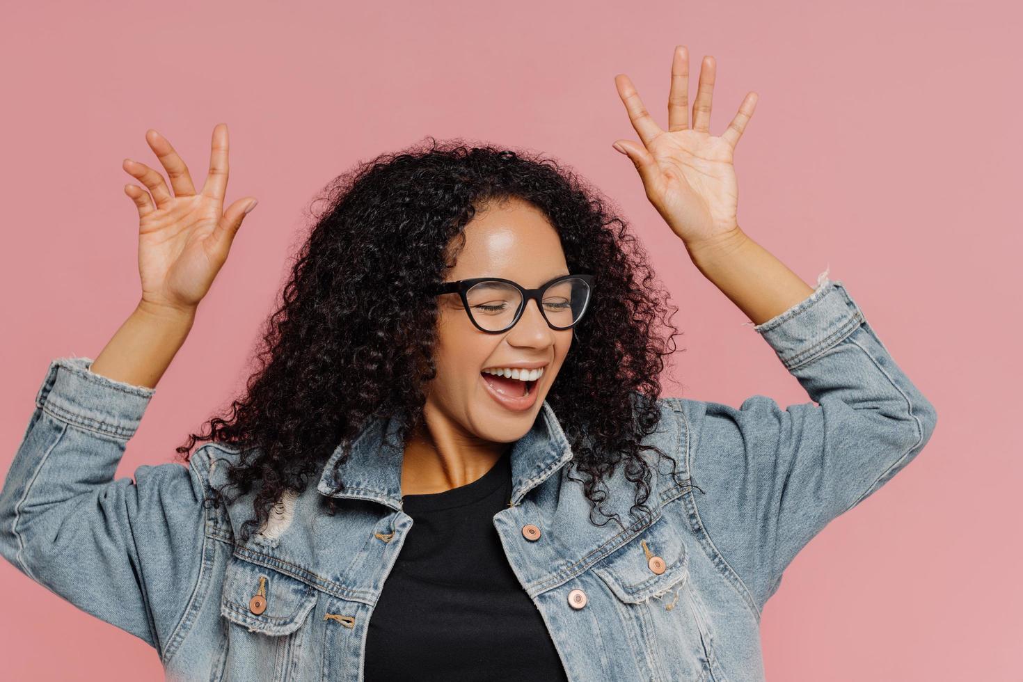 Carefree overjoyed dark skinned young woman dances from happiness, has fun indoor, keeps arms raised, closes eyes in pleasure, wears spectacles and denim jacket, isolated on pink background. photo