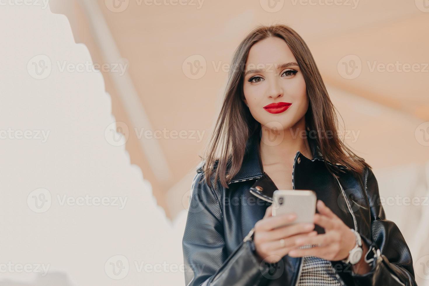 Horizontal shot of attractive dark haired female with red painted lips, dressed in black leather jacket, holds modern cell phone, sends text messages, chats online, strolls at city street. Technology photo