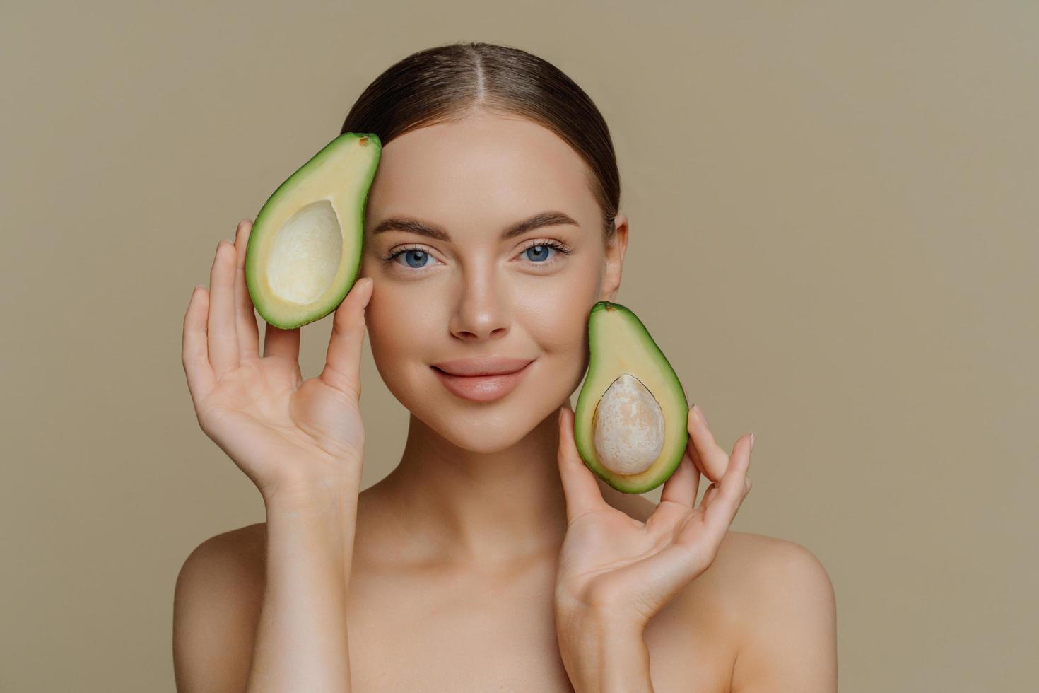 Photo of shirtless female model looks with gentle expression at camera holds halves of avocado near face has healthy well cared skin after cosmetic treatments isolated over beige background.