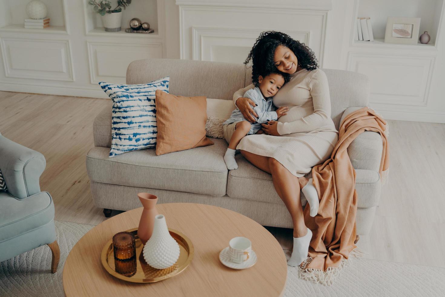 Single african pregnant mom tenderly embracing little son while sitting on cozy couch photo
