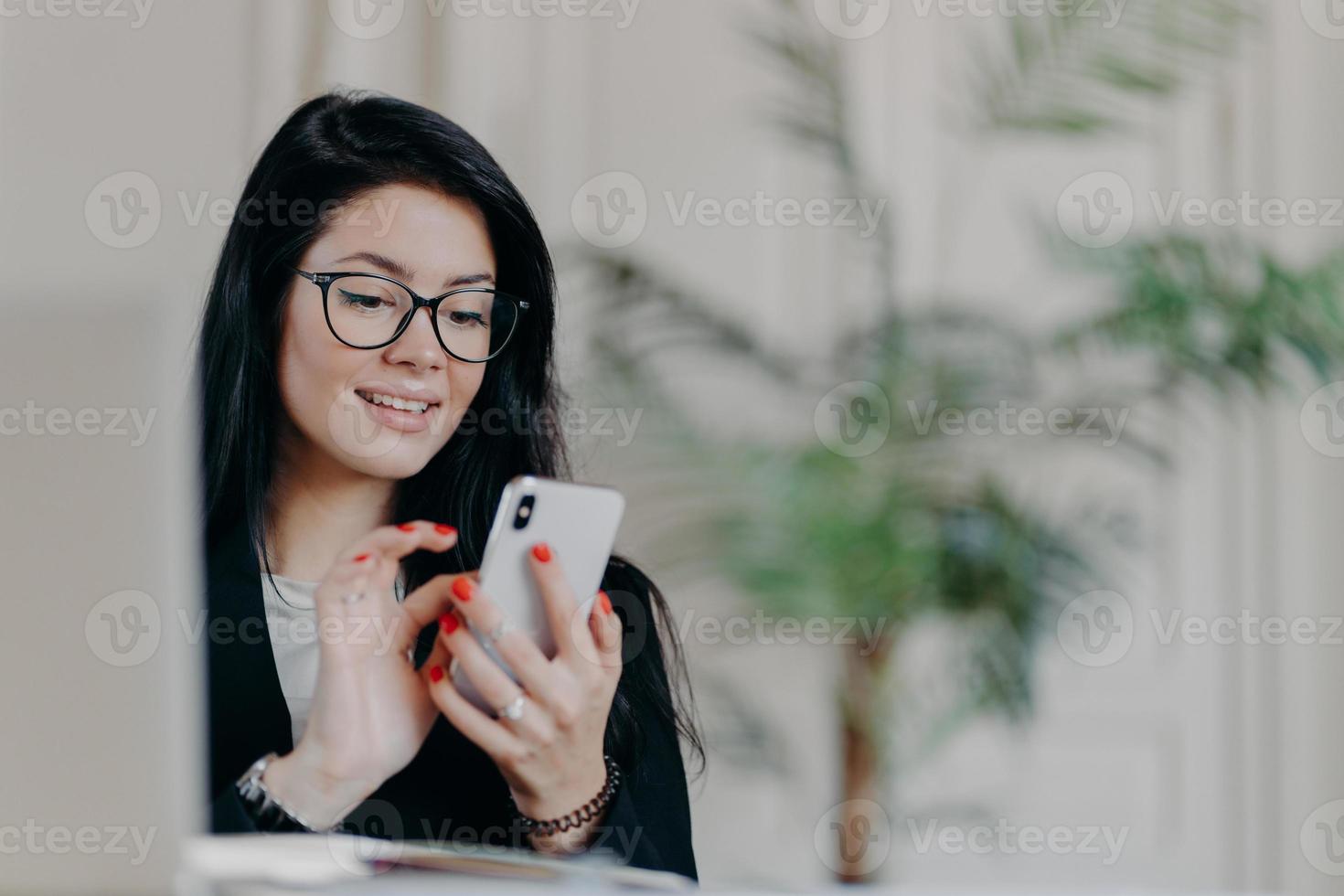 Happy brunette businesswoman uses smartphone, wears spectacles, sends text messages to partner, works on project, poses against cozy interior at workplace. Technology, occupation and work concept photo