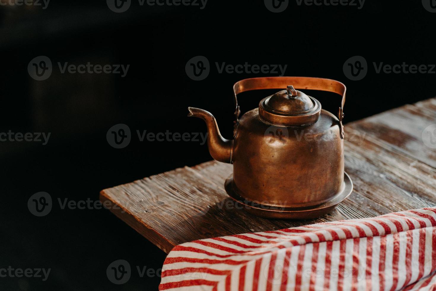 Horizontal shot of antique kettle on vintage wooden table. Old crockery. Metal copper teapot against dark background red towel near. Selective focus photo