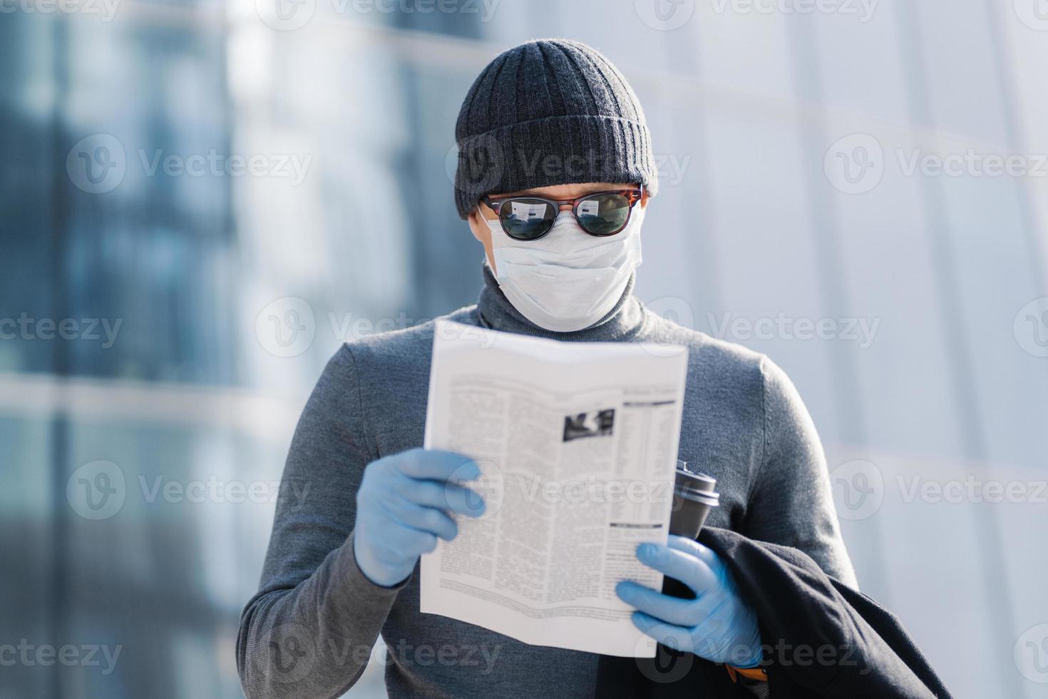 Shot of young man reads article about coronavirus symptoms, wears protective medical mask and rubber gloves, poses at street with coffee, follows rules or quarantine. Covid-19 protection concept photo
