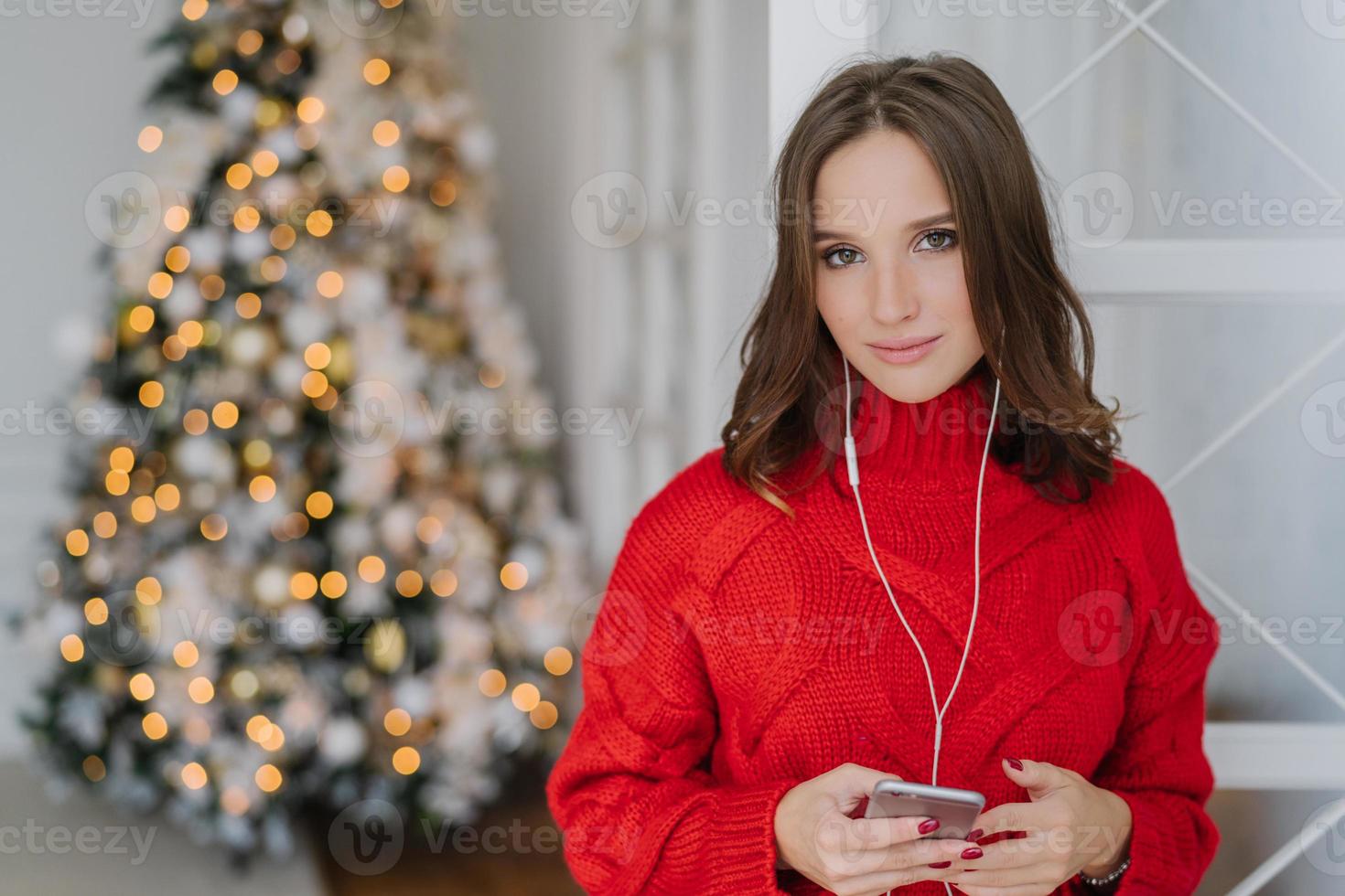 Photo of cute young woman with dark hair, dressed in knitted red sweater, holds smart phone in hands, listens radio online, connected to wifi at home, pose near beautiful New Year tree with lights