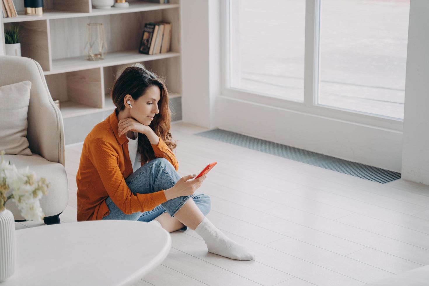 Spanish girl is sitting on floor at home near window in morning and listening to music on telephone. photo