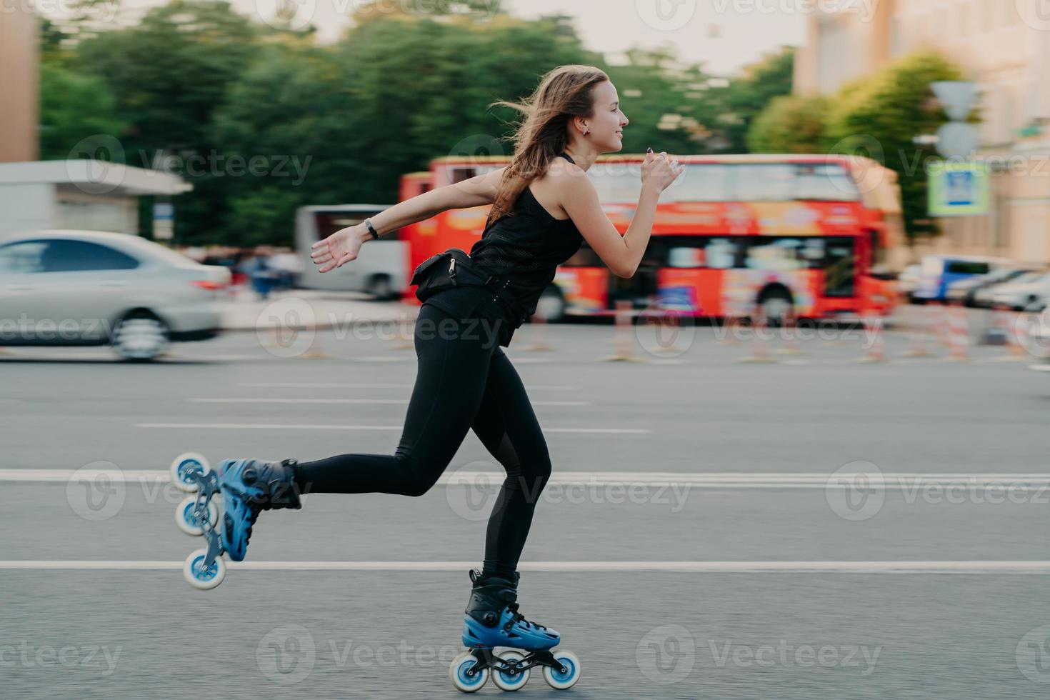 Young fit woman on roller skates with wheels rollerblades during summer day on busy road with transport leads active lifestyle wears black sportsclothes breathes fresh air. Movement concept. photo