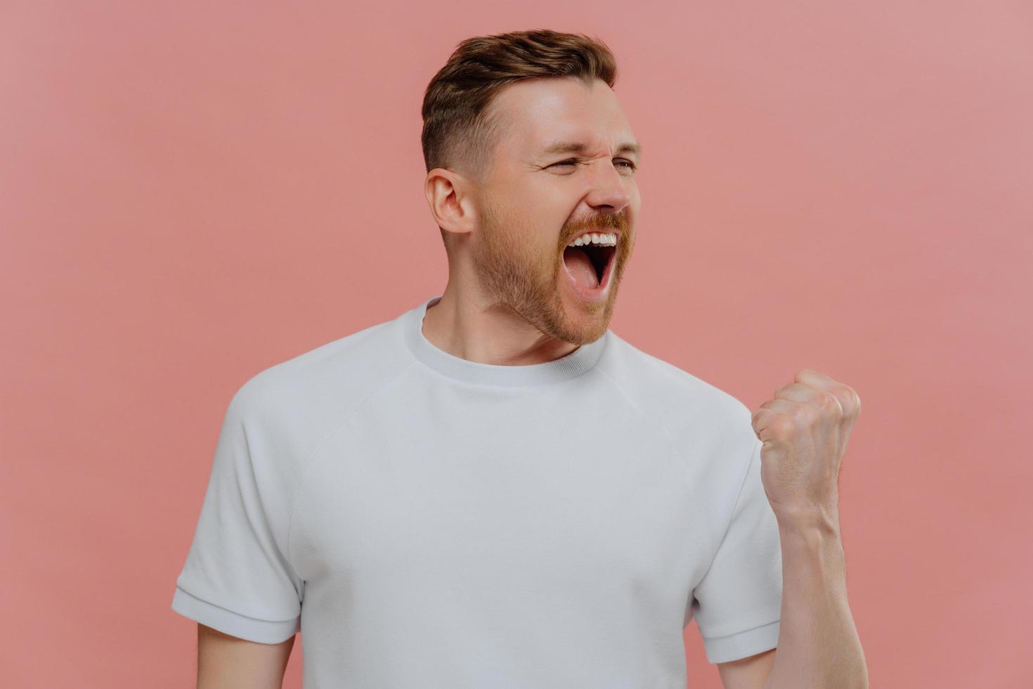 Emotional successful man shouts from joy clenches fist with triumph makes pump gesture cellebrates triumph dressed in casual white t shirt isolated over pink background. Body language concept photo