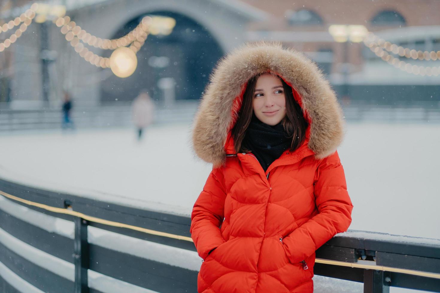 Image of lovely thoughtful young woman wears winter red coat with fur on hoody, keeps hands in pockets, stands against blurred background near skating rink, has outdoor walk during winter time photo