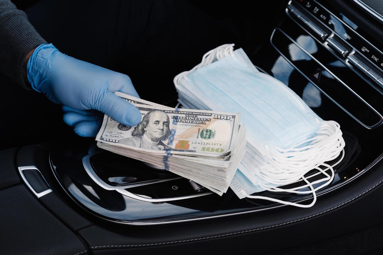 Virus concept. Hand in rubber gloves holds stack of dollar banknotes, earns money as sells medical masks, poses in car. Covid epidemic. Speculation at market of necessary products. Quarantine period photo