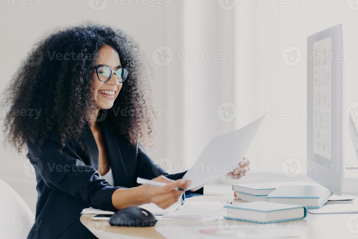 Glad Afro American woman with curly hairstyle, makes financial report, holds paper documents, has happy smile wears spectacles for vision correction dressed in elegant clothes computer screen in front photo