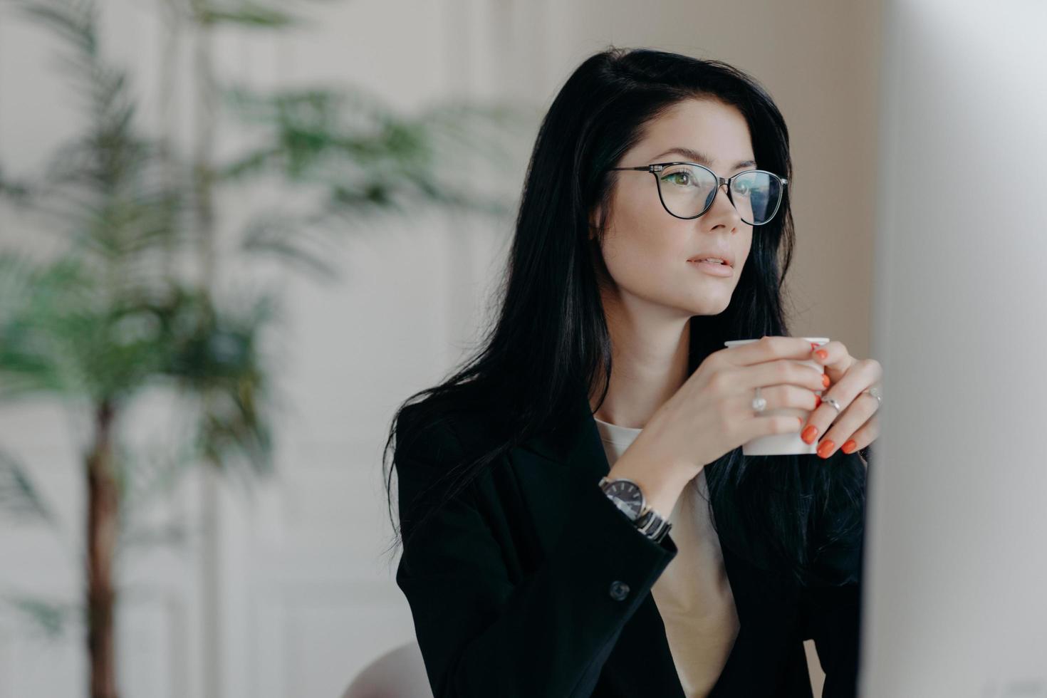 Photo of concentrated brunette female copywriter drinks coffee, works on creative task, wears spectacles and black formal costume, watches webinar to improve skills, poses in coworking space