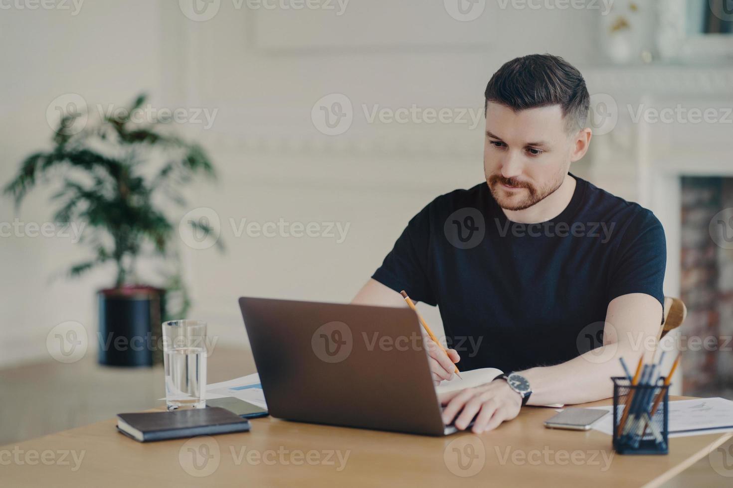 Serious bearded man writes informative notes while watching webinar on laptop computer works remotely dressed in casual t shirt poses at workplace against home office interior. Modern workspace photo