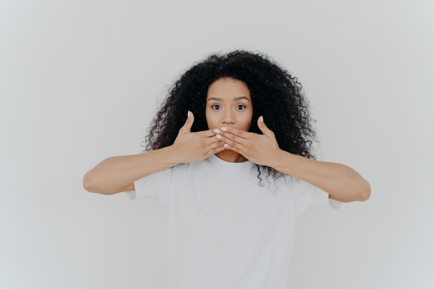 toma horizontal de una mujer sorprendida de piel oscura que cubre la boca con ambas palmas, trata de no hablar, vestida con una camiseta casual de maqueta, posa contra un fondo blanco. gente, concepto de secreto. foto