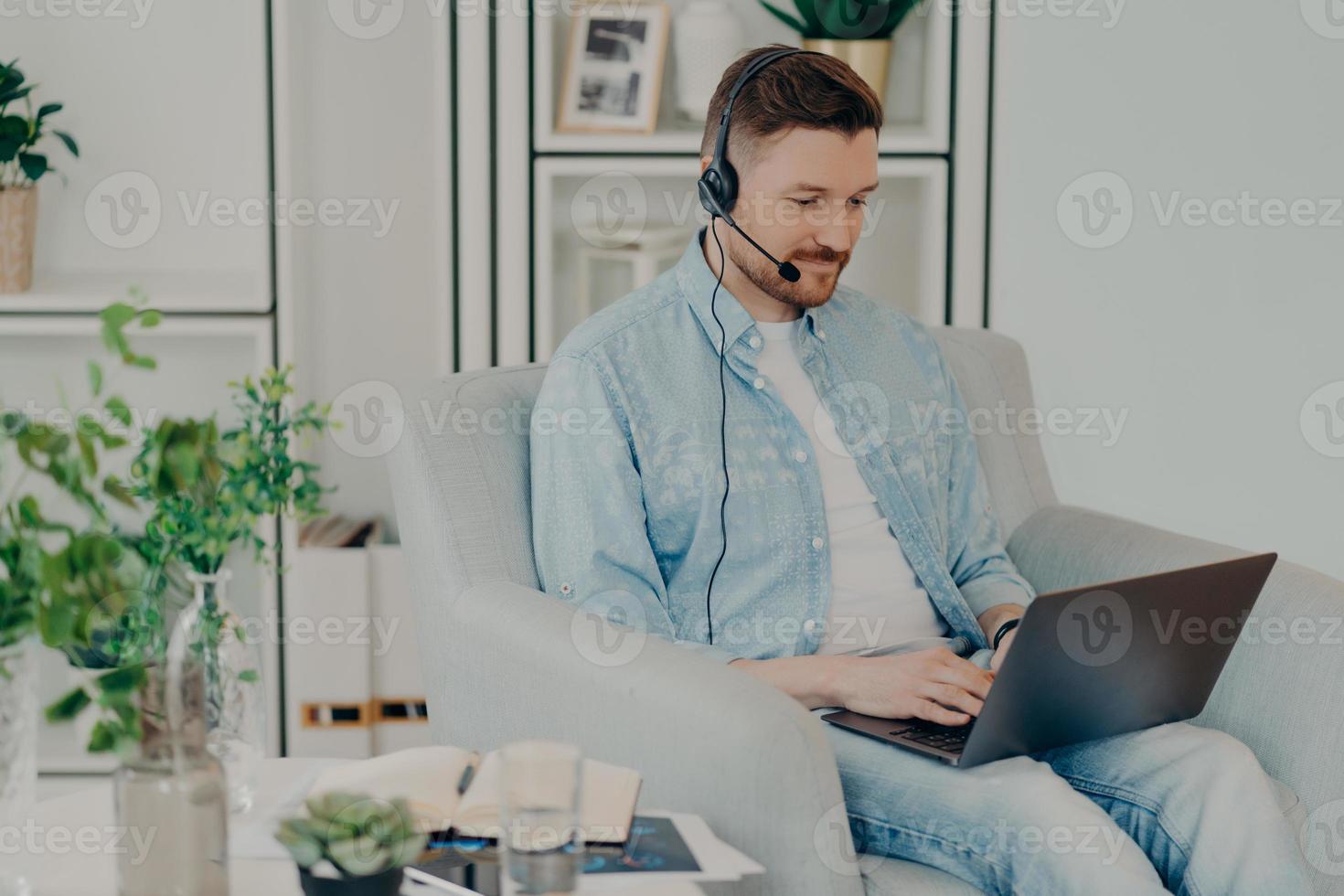 Busy man dressed in denim clothes supplies consultant service types on laptop computer wears headphones with microphone works distantly sits in comfortable armchair participates in online negotiations photo